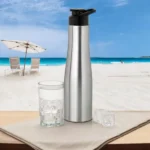 Portable stainless steel water bottle displayed beside glass on beach setting.